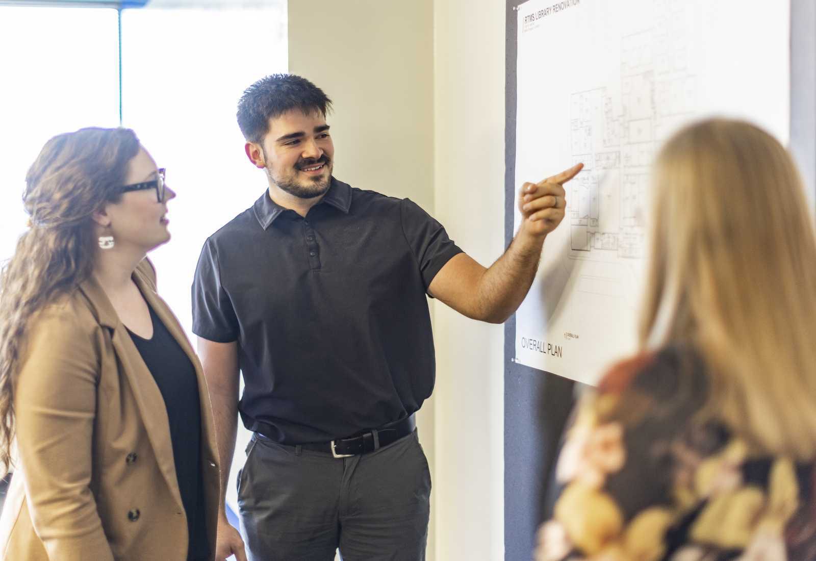 A man points at a floor plan while discussing ideas with two women in a modern office setting. Everyone appears engaged and attentive.
