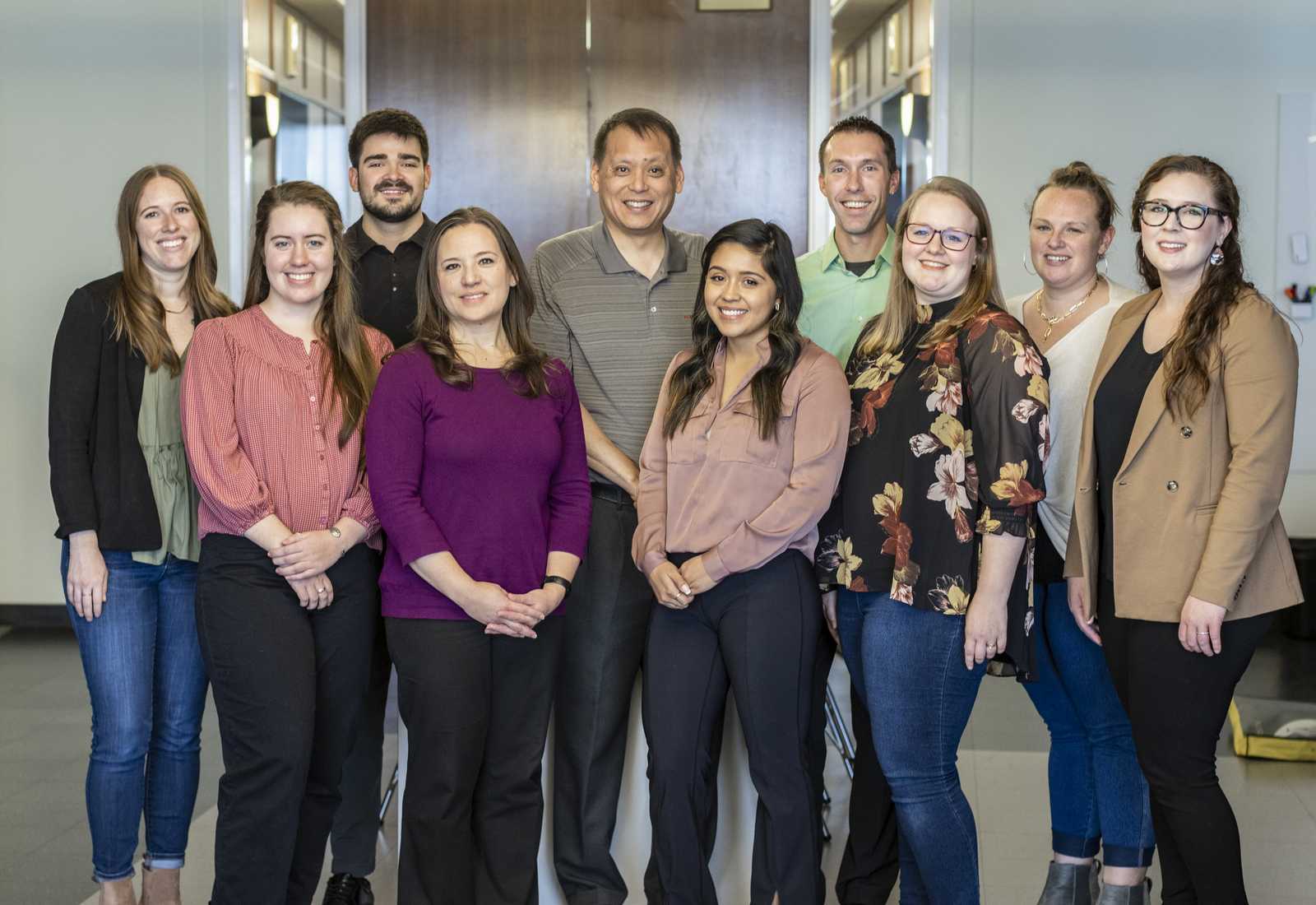 A group of ten individuals, smiling and posing together in a bright office setting, showcasing a diverse range of clothing styles.