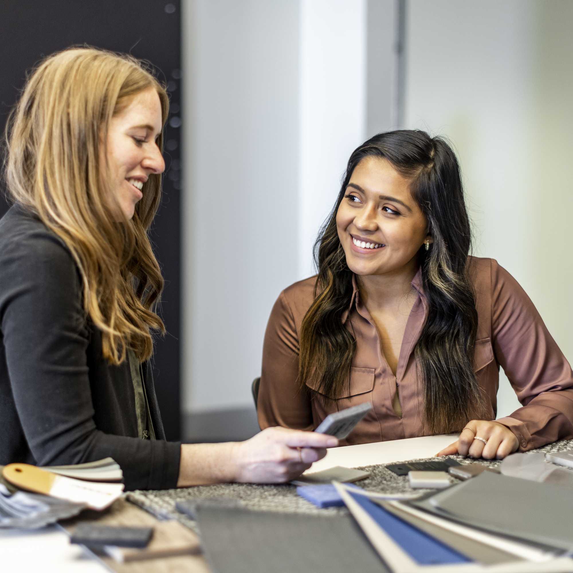Two women share a friendly conversation while reviewing design materials at a table, displaying collaboration and creativity.