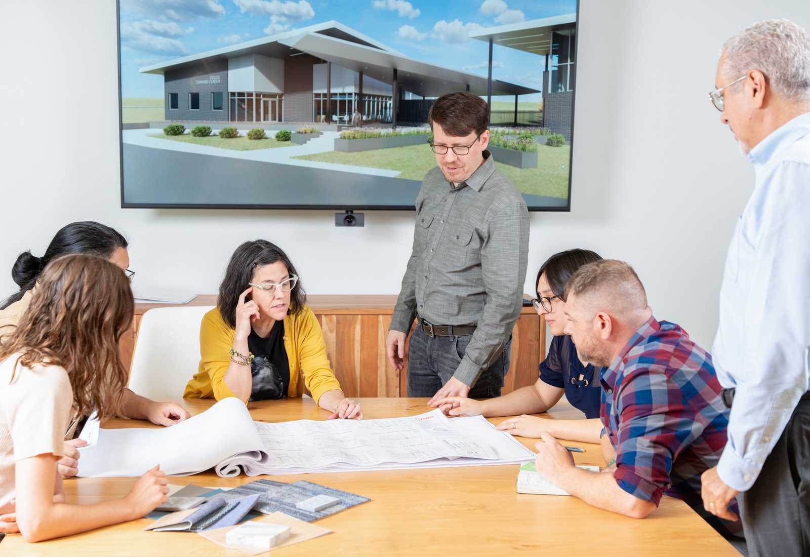 A group collaborates around a table, reviewing architectural plans, with a digital rendering of a building displayed on the screen behind them.