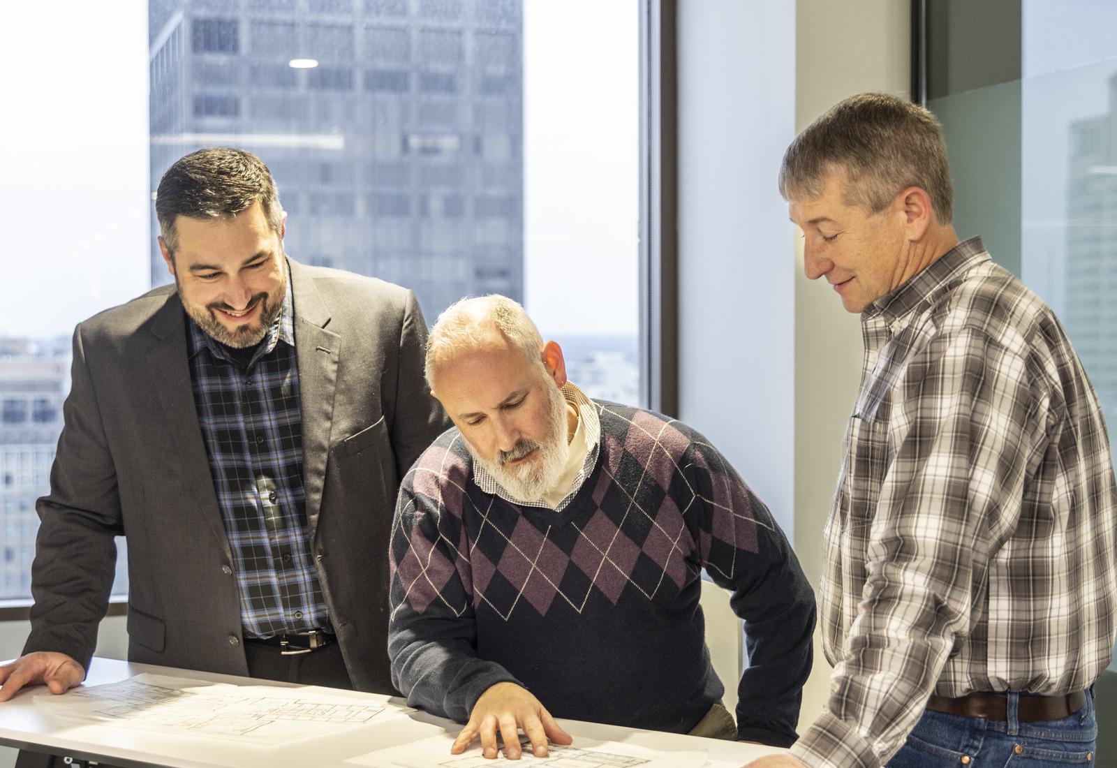 Three men are discussing architectural plans while standing around a table, with a cityscape visible through large windows in the background.