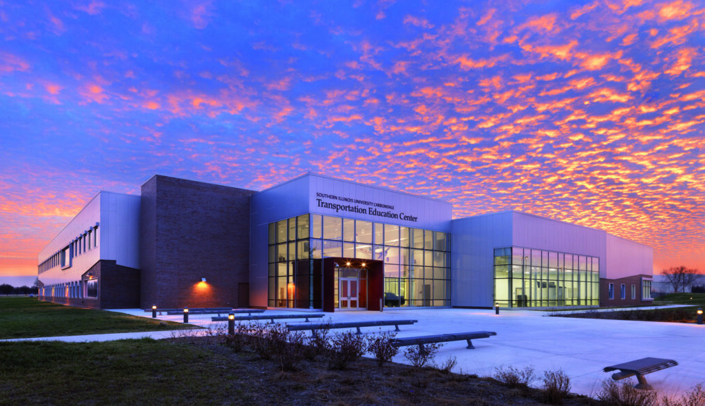 A modern educational building with large glass windows stands against a vibrant sunset, highlighting its contemporary architecture and surrounding greenery.