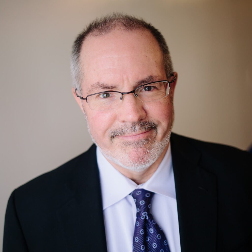 A smiling man with glasses and a light beard wears a dark suit and tie, exuding professionalism and approachability.