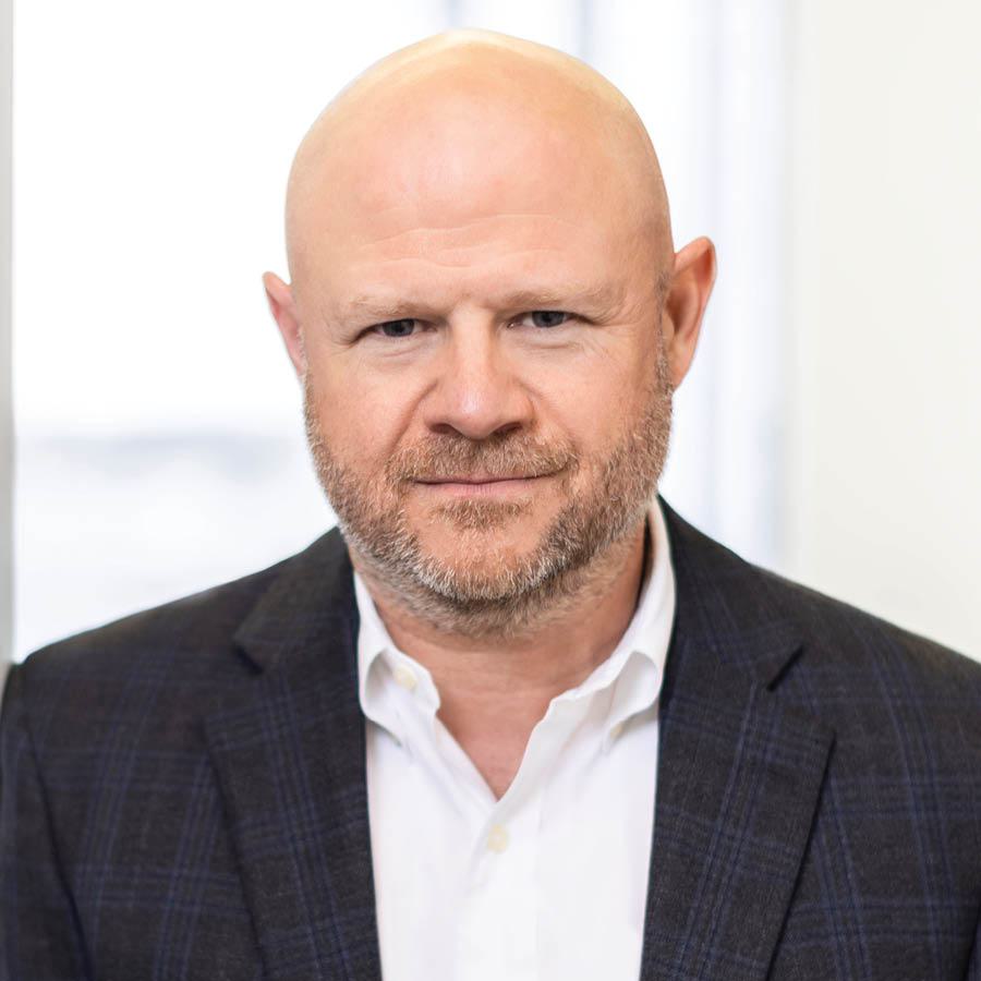 A confident man with a bald head and a beard wears a dark blazer over a white shirt, standing against a bright, blurred background.