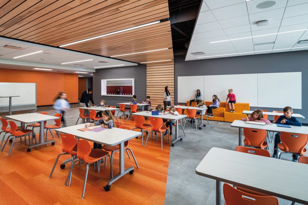 A modern classroom features colorful seating and various activity areas, with students engaged in different tasks and teachers supervising.