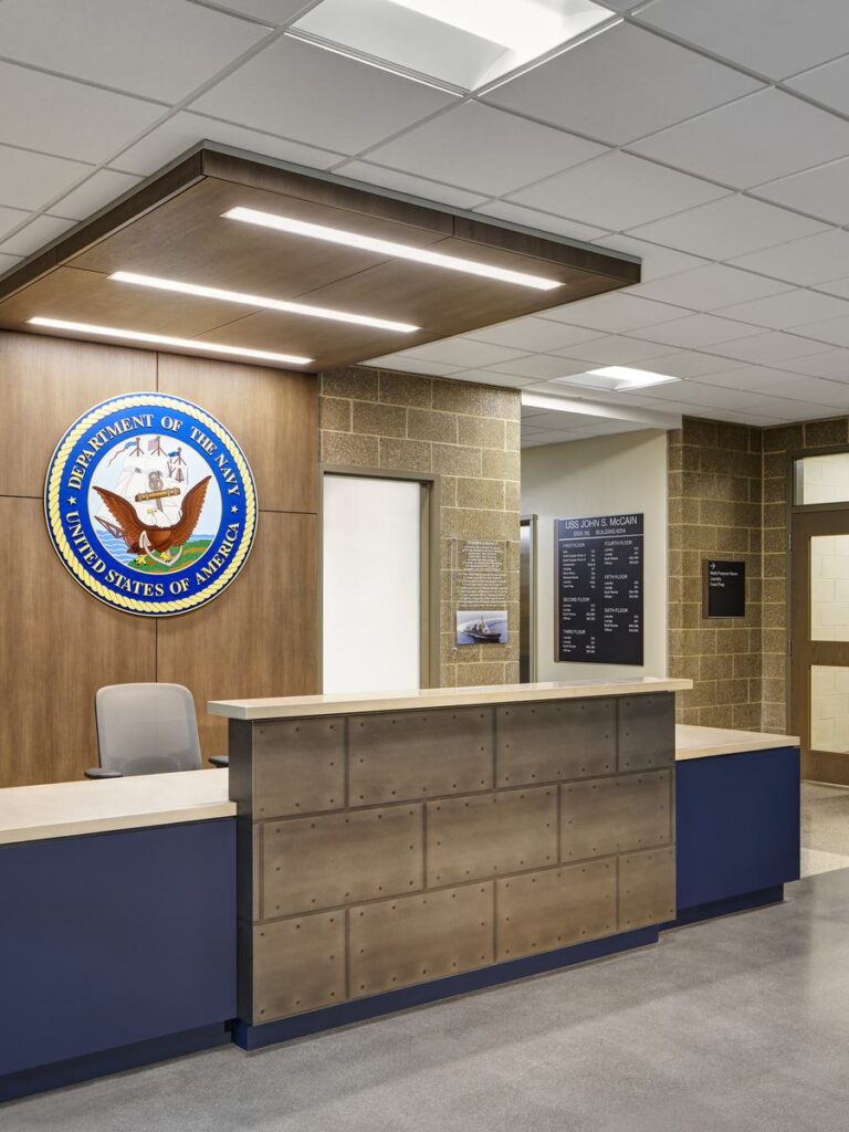 A modern reception area features a wooden front desk, a prominent seal of the Department of the Navy, and neutral, well-lit decor.