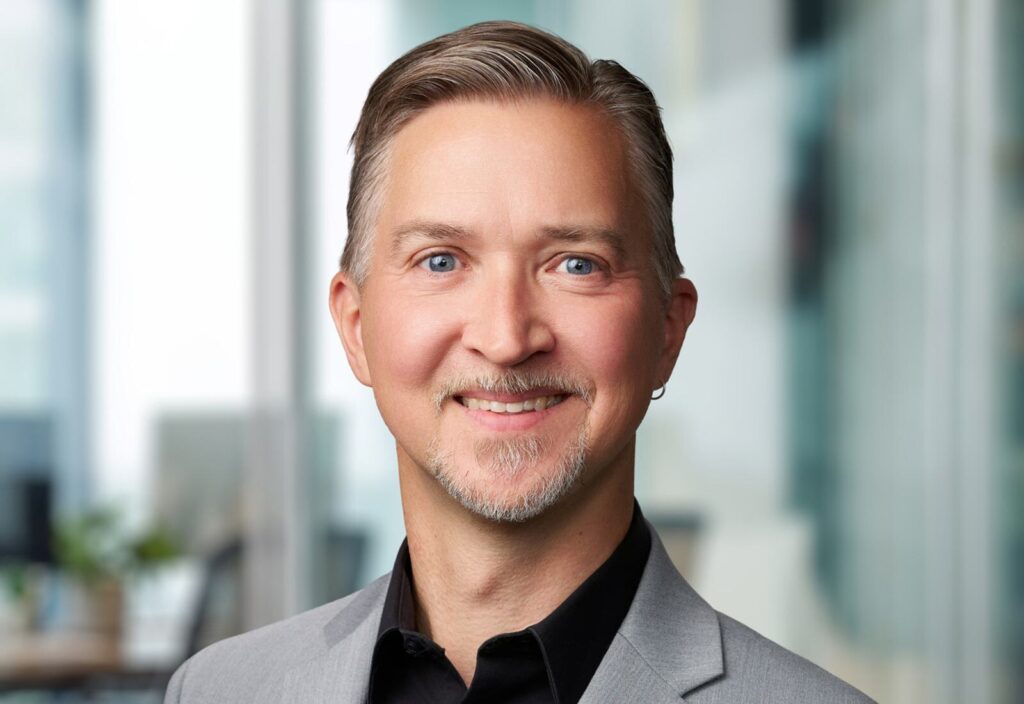 A smiling individual with light gray hair and a neatly trimmed beard wears a gray blazer over a black shirt, set against a blurred office background.