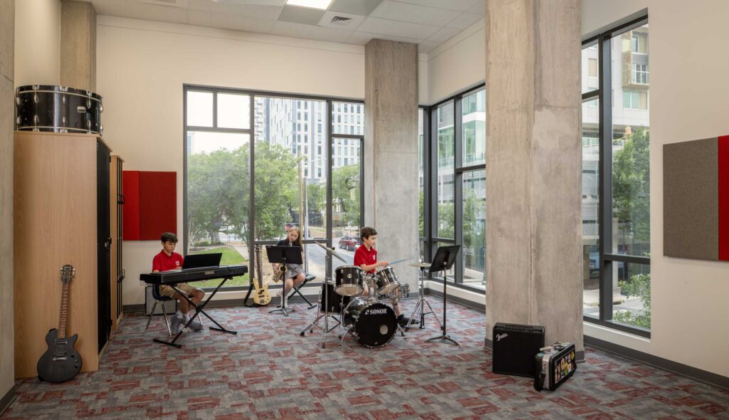 Three children are rehearsing music with a keyboard, drums, and guitar in a bright studio featuring large windows and modern decor.