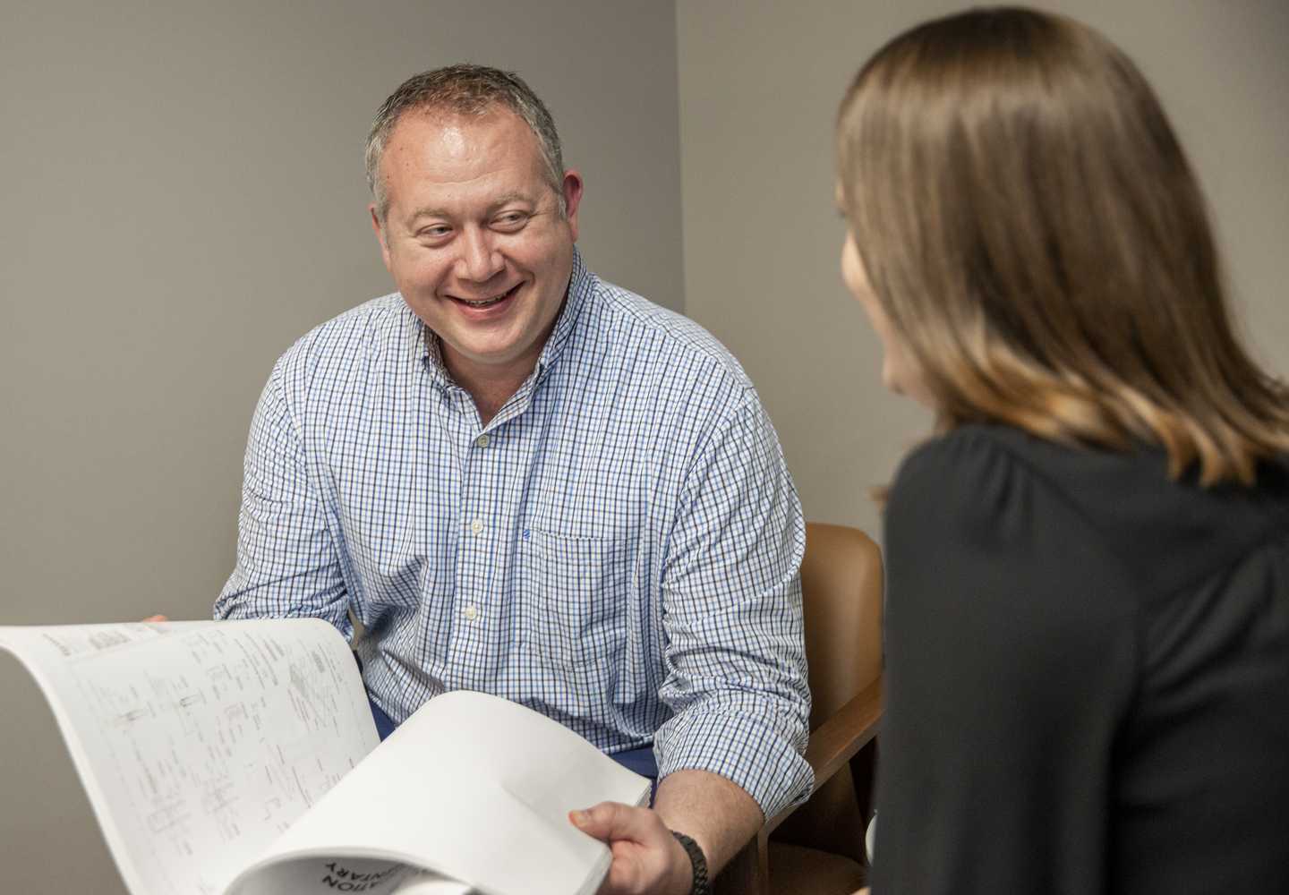 A man in a checked shirt smiles while discussing documents with a woman, fostering a collaborative and engaged conversation.
