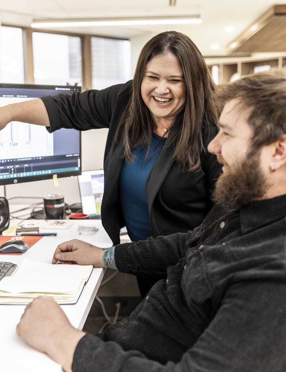 Two colleagues share a lighthearted moment in an office, discussing work on a computer screen, with a collaborative and engaging atmosphere.
