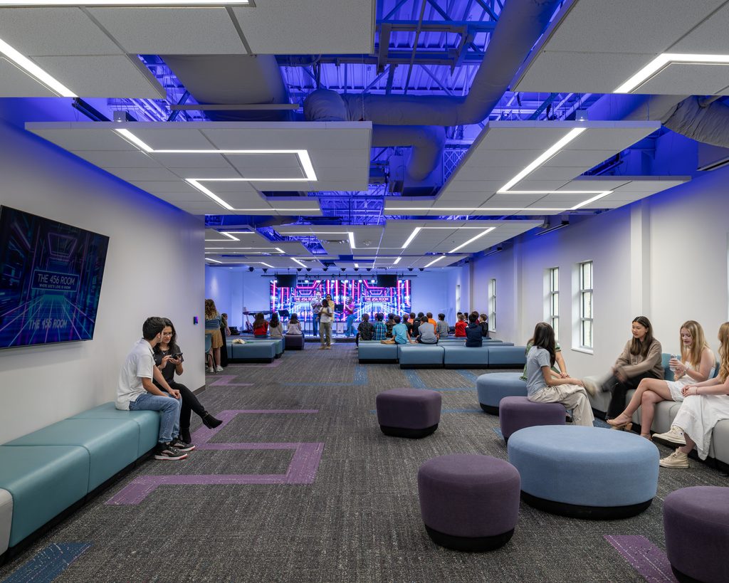 Church youth room with teens/young adults gathered in various flexible spaces on movable blue and purple furniture. Design of the room reads "tech" with angular lines and blue and purple technology-inspired graphics.
