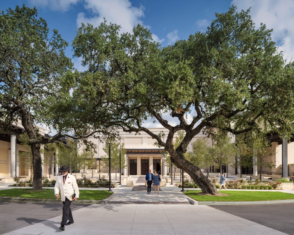 Exterior courtyard of a church campus featuring mature oak trees, landscaped gardens, wide pedestrian walkways and light-colored brick buildings, with visitors walking toward the main entrance.