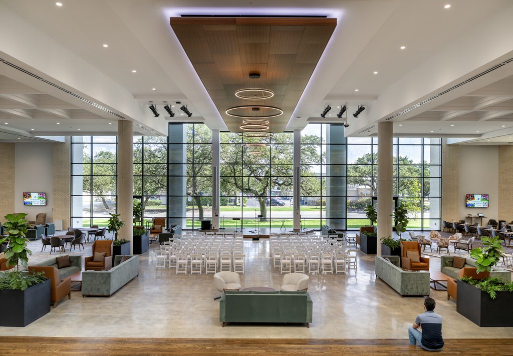 A large banquet hall with expansive floor-to-ceilikng windows, tall ceilings, and neutral-colored furniture. One singular man sits on the steps leading down to this space, in the lower right corner, a small piece of a much grander space.