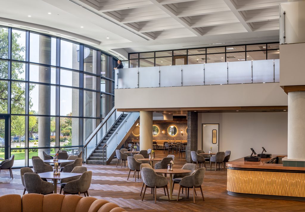 Banquet hall or grand lobby area with curved floor-to-ceiling windows on the left, neutral shaded tables and couches in the foreground, restaurant seating and a cafe counter in the background, and a second floor mezzanine with a staircase to access.