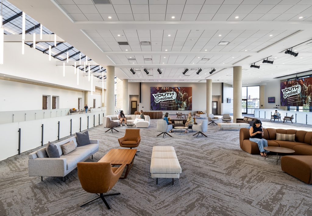 A modern, fresh church youth room that opens on one side to a multi-story atrium with glass ceiling windows. Neutral brown, grey and cream furniture and bold carpet accents the bright room.