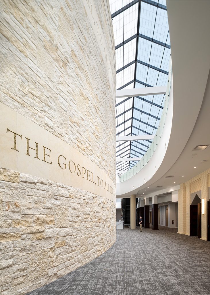 Long shot of a hallway in a two-floor atrium with a curved cream-colored stone wall on the left that spans both stories, natural light shining through the pitched full-glass ceiling, and a man near the lower middle-right for scale (he is very small in this grand image).