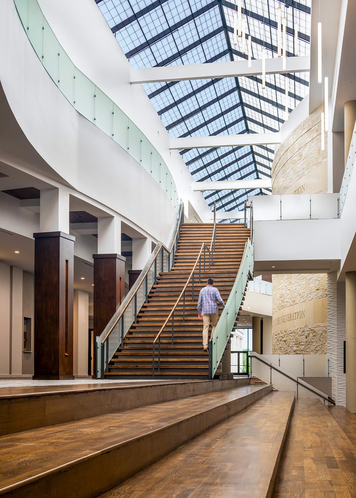 Man walks up a sleek, clean-lined wooden staircase in a multi-story atrium with abundant natural light flooding from above.