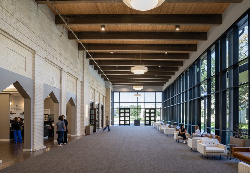 Spacious church lobby with white brick walls, exposed wood ceiling beams, floor-to-ceiling windows and lounge seating, with visitors gathering near a café.