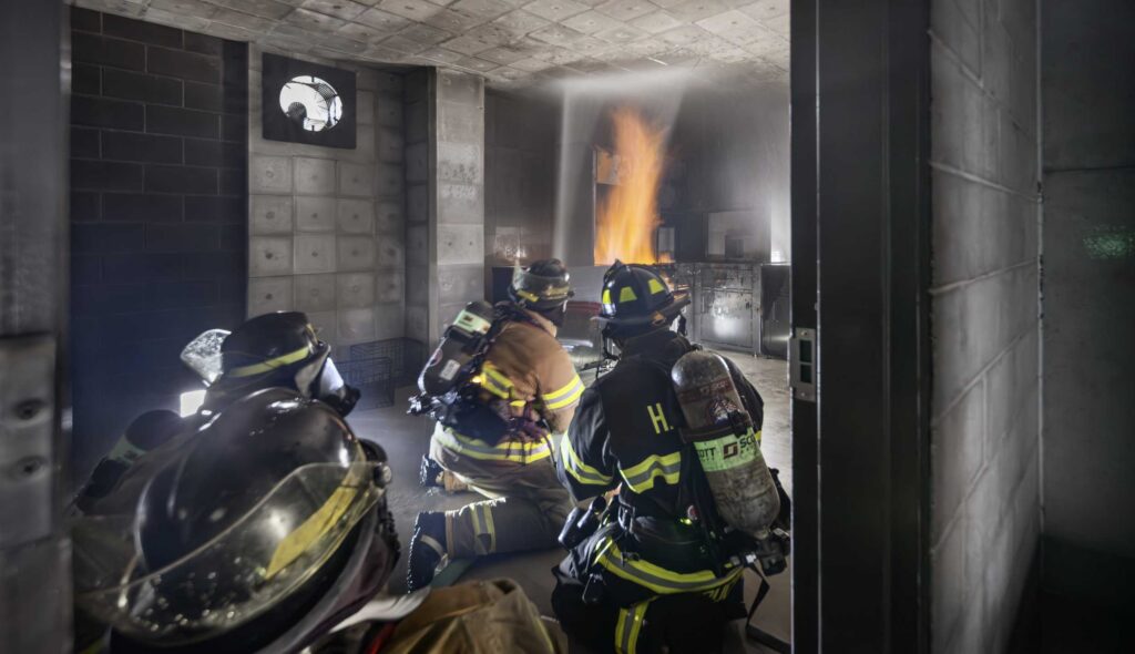 Four firefighters in full gear inside a training room dousing a live fire in the Harper College Emergency Services Training Center.