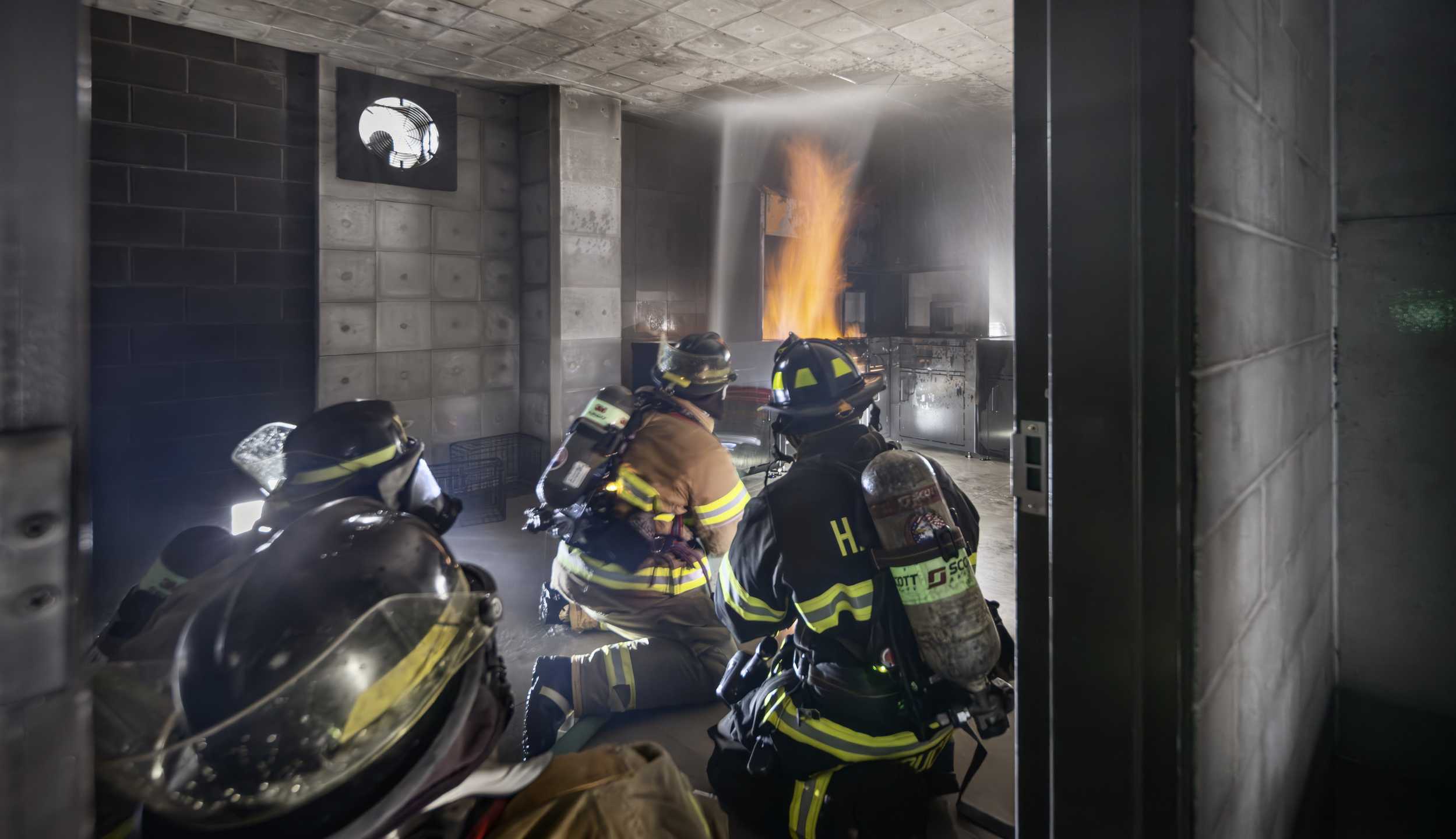 Four firefighters in full gear inside a training room dousing a live fire in the Harper College Emergency Services Training Center.
