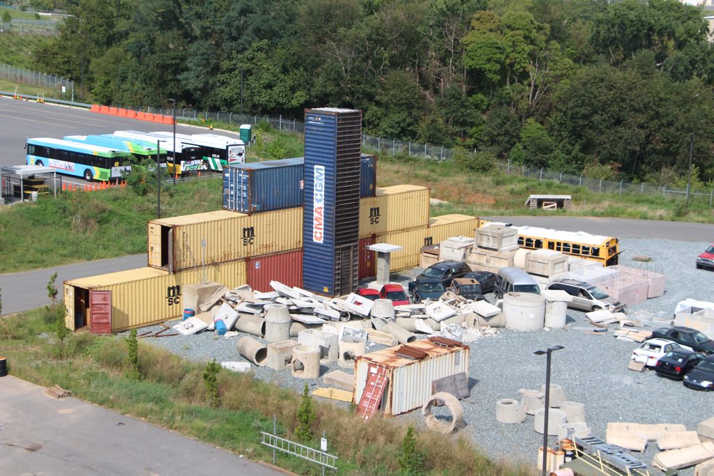 Outdoor junk yard-esque training area for first responders, including piled cars, construction materials, concrete pieces, shipping containers, a school bus and more