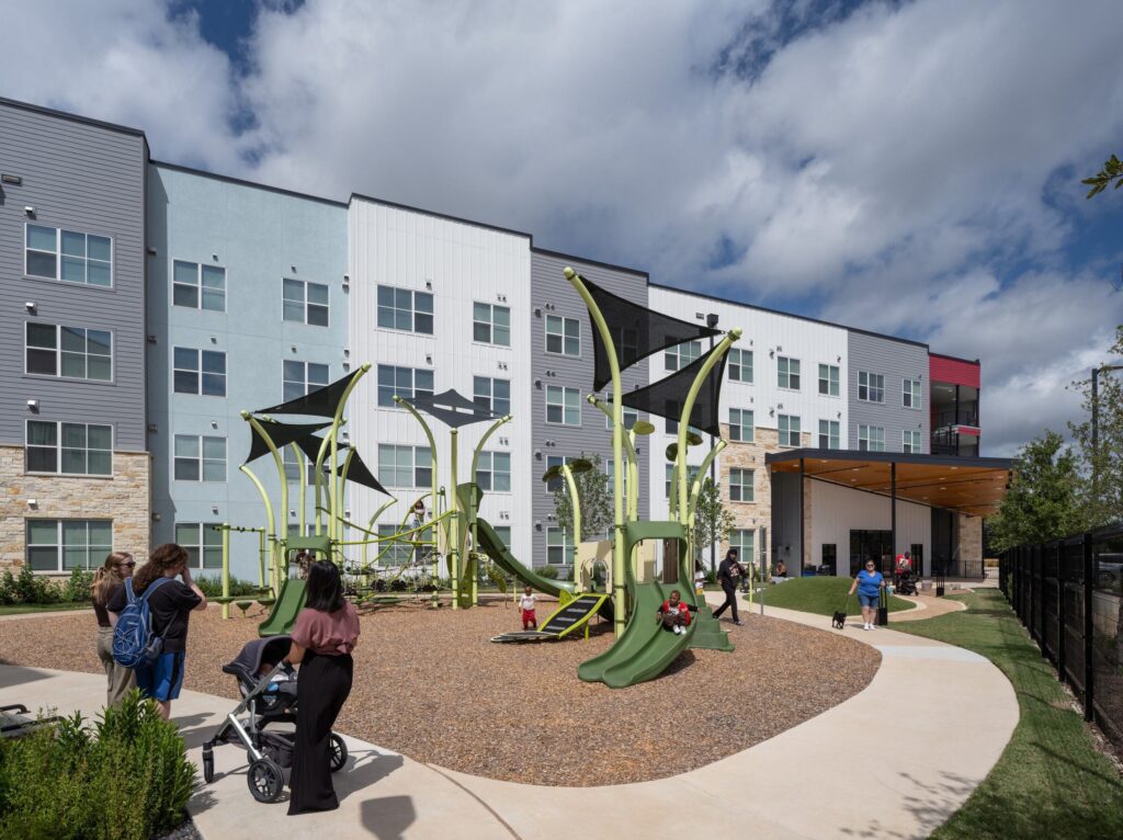 A lively playground with unique green structures sits in front of colorful apartment buildings, as families enjoy the outdoor space.