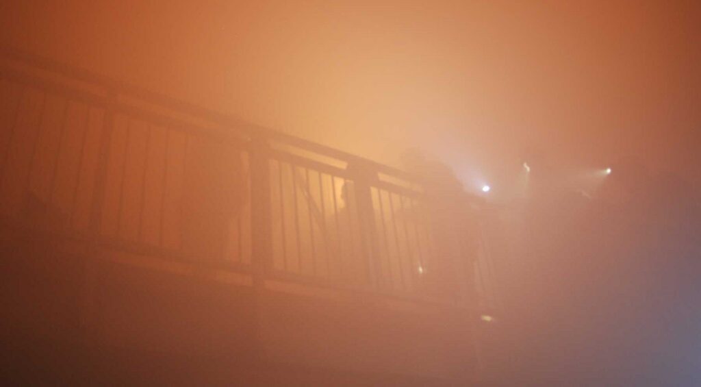 A dense fog engulfs a railing, silhouettes of people using flashlights emerge, creating a mysterious and atmospheric scene.