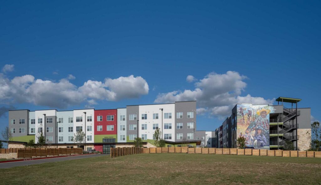 Wide exterior view of the Norman Commons apartment community on a clear day, featuring a modern four-story residential building with white, gray and red façade panels, large windows and green accents. A second building connected by a central walkway displays a large colorful mural on its end wall. A grassy open space and wood fencing frame the development beneath a bright blue sky with scattered clouds.