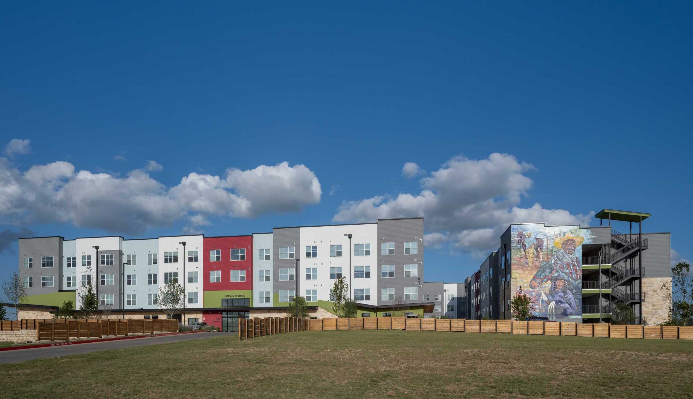 Wide exterior view of the Norman Commons apartment community on a clear day, featuring a modern four-story residential building with white, gray and red façade panels, large windows and green accents. A second building connected by a central walkway displays a large colorful mural on its end wall. A grassy open space and wood fencing frame the development beneath a bright blue sky with scattered clouds.